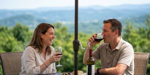 Couple enjoying a relaxed outdoor wine tasting at a Hudson Valley winery, seated at a patio table with vineyard views and rolling hills in the background.